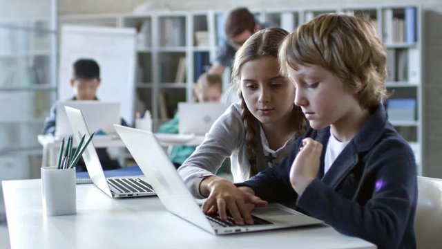 Girl of primary school age helping her classmate with task that they are doing on laptop computer