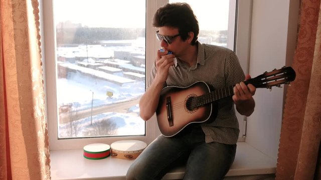 Playing a musical instrument. Man is playing the mouth organ sitting in windowsill