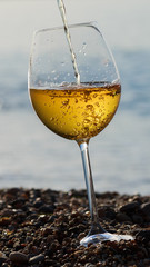 Alcoholic beverage of white wine pouring into glass outdoor on summer day on blue sea.