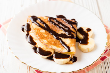 Crepes with banana and chocolate syrup on white wooden background.