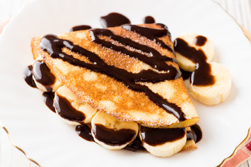 Crepes with banana and chocolate syrup on white wooden background.