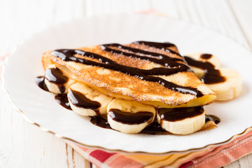 Crepes with banana and chocolate syrup on white wooden background.