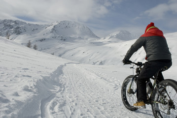 man use electric bicycle, e-bike, ebike, pedal on snow covered road, downhill mountain, specific bike with wide wheels to go on snow, called fatbike, winter, cold, alps, Simplon Pass, Switzerland