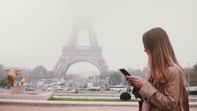 Portrait of young happy woman standing near the Eiffel tower in Paris, France and using the smartphone. Slow motion.