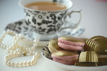 Gold and pink French macarons with pearls and a fine bone china cup with black flowers. Luxurious treats. 
