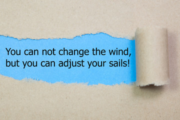 Motivational quote You can not change the wind but you can adjust your sails, appearing behind torn paper.