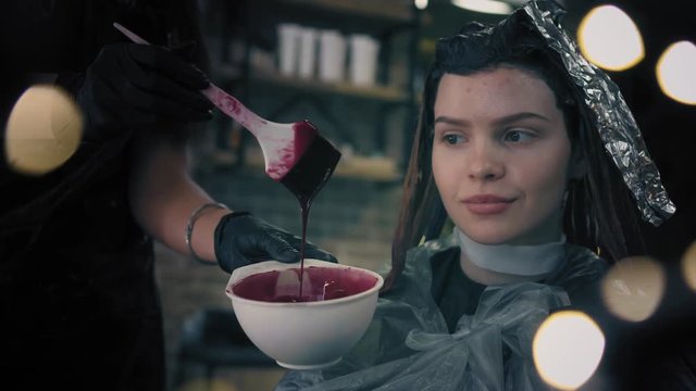 Hairdresser is mixing red hair dye in white plastic bowl with brush. Female is sitting on chair with fully dyed hair in hair salon and watching the process
