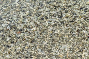 Sea pebbles under the water. Transparent sea water. Through the clear water can see the seabed. Natural colors and a natural texture for the background. Clear sea water with patches of sunlight.
