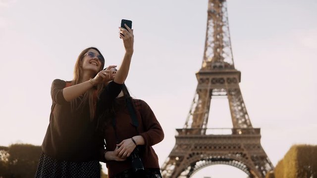 Two beautiful traveling female taking photos on smartphone near the Eiffel tower in Paris, France in sunny day.