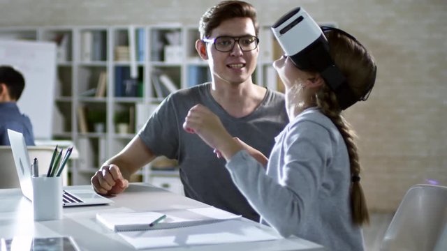 Girl of primary school age sitting in VR glasses and learning to use virtual reality with her teacher during computer coding class