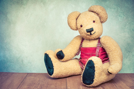 Retro Teddy Bear toy dressed in red leather clothes sitting on wooden floor front textured concrete wall background. Vintage style filtered photo