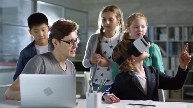 Group of four primary school children, one of them wearing VR glasses, learning to use virtual reality with their teacher