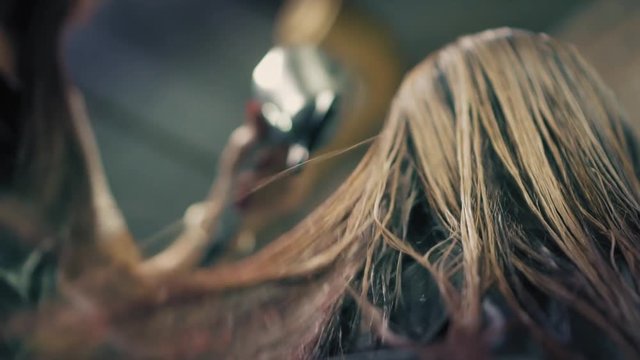 Woman with long blonde hair is sitting in hair salon while hairdresser is drying her wet hair with hair dryer and hairbrush, Closeup, down back angle. Slow mo.