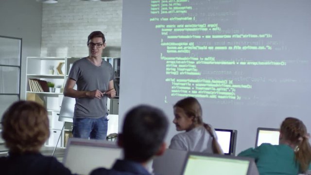 Young teacher explaining topic to primary school children with the help of projector in computer coding class, students participate actively and raise their hands during lesson