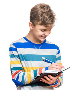 Portrait Of Caucasian Smiling Teen Boy With Notebook And Blue Pen Writing Something. Handsome Funny Teenager, Isolated On White Background. Happy Student Writing On Note Pad.