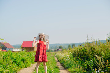 little caucasian blonde girl in red dress and white hat with large brim posing on dirt road in countryside background