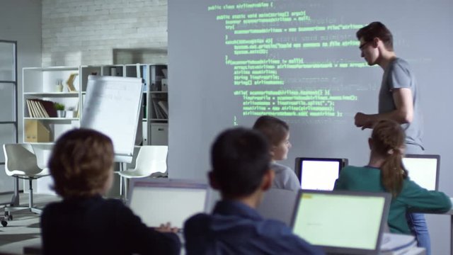 Young teacher walking in class and explaining topic to primary school children with the help of projector during computer coding lesson