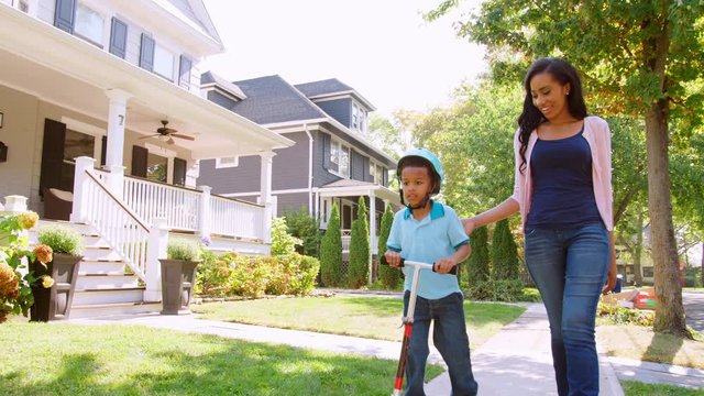 Mother Walks With Son As He Rides Scooter Along Sidewalk