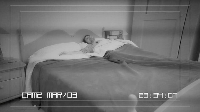 A CCTV camera screen: a sleeping man's reaction to something scary or annoying happening in his bedroom.
