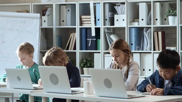 Tracking shot of four primary school children doing written task during information technology class and looking at flip chart schemes left by their teacher