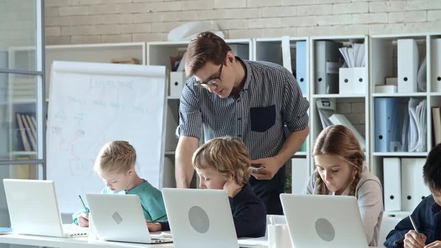 Four primary school children writing something in copybooks during information technology class, their teacher helping one of them