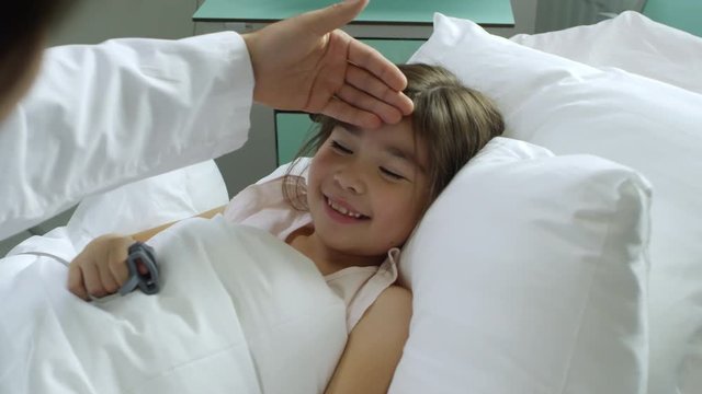 Medium shot of male pediatrician putting hand to forehead of smiling girl lying in hospital bed and checking her temperature