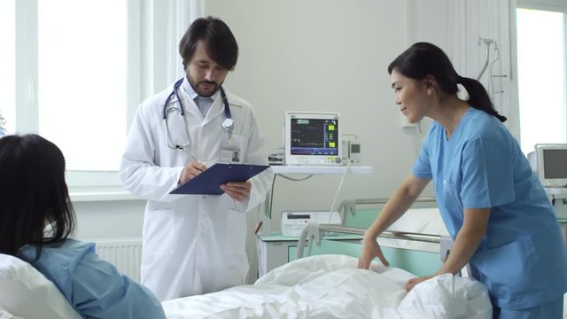 PAN of unrecognizable woman lying in hospital bed and shaking hand of bearded male doctor in lab coat filling patients form on clipboard