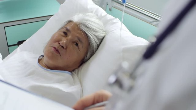 Medium shot of cheerful elderly woman lying in hospital bed and talking with unrecognizable doctor making notes of clipboard