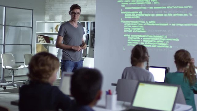 Young teacher explaining topic to primary school children with the help of projector in coding computer class