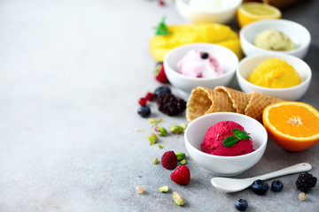 Red, pink, yellow, green, white ice cream balls in bowls, waffle cones, berries, orange, mango, lemon, mint, pistachio, grey concrete background. Ice-cream colorful collection, summer concept