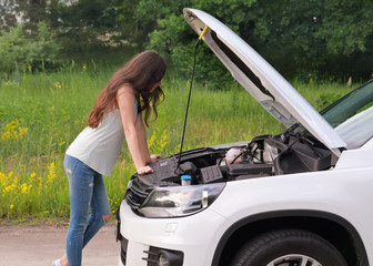 Girl examining her broken car on the country road. Young brunette woman trying to repair her broken car with opened hood.