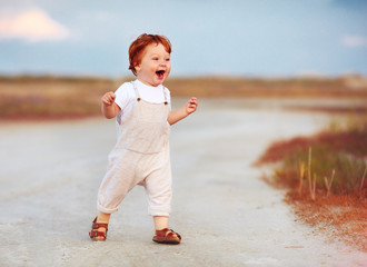 adorable redhead toddler baby boy in jumpsuit running through the summer road and field