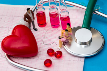 small toy people are on a cardiogram near a stethoscope, heart and tablets on a blue background