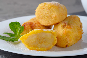 Bananas in batter and sprinkled with powdered sugar on a white plate on the gray slate background. Close-up view.