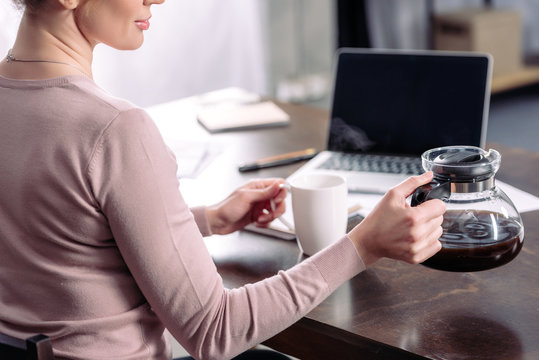 Partial View Of Woman With Cup And Coffee Maker Sitting At Table With Laptop At Home