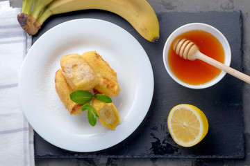 Bananas in batter and sprinkled with powdered sugar on a white plate on the gray slate background. Top view.
