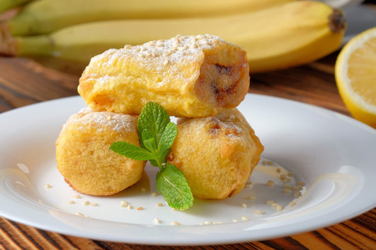 Bananas in batter and sprinkled with powdered sugar on a white plate. Fruits on the wooden background. Close-up view.