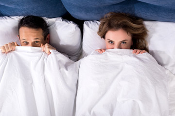 overhead view of wife and husband hiding under blanket while lying in bed at home