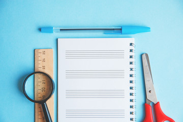 pen, ruler, magnifier and scissors on a blue background. Education or business concept. Empty white note place for text.