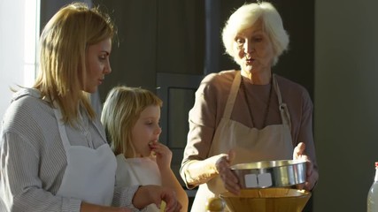 Joyous women cooking at home together: young woman cutting apple with little girl while grandmother sifting flour at kitchen counter