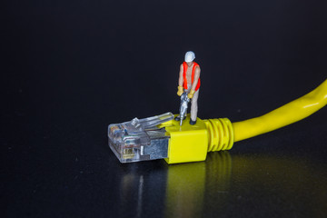 Miniature workman using a hammer drill on a yellow network cable and plug over a black background with copy space in a conceptual image