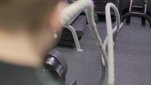 Young guy is in intensive training with rope in sports club. Male athlete pulls strong cord in modern gym indoors. Bodybuilder stands and holds rope, moves with powerful hands actively, creating