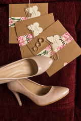 Wedding invitations in the craft envelopes, golden rings and a beige bridal shoes lying on a red armchair. Wedding concept. Wedding accessories. 