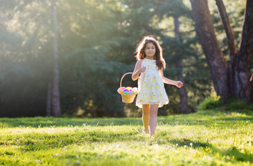 Little girl walking on green grass toward camera holding woven basket with pastel Easter eggs