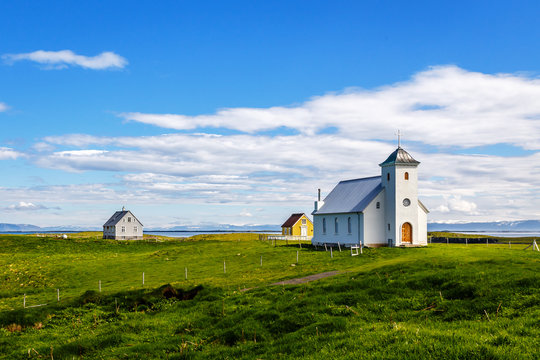 Flateyjarkirkja church and couple of living houses with meadow in foreground and sea  fjord with blue sky in the background, Flatey, Iceland