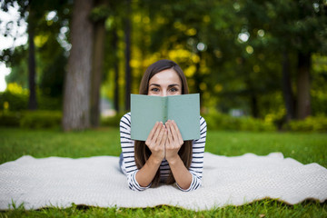 Beautiful smiling modern young girl student in casual clothes reading a book lying on a plaid in a...