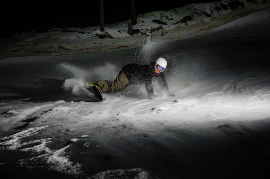 Young and active snowboarder riding down the snowy mountain slope at night