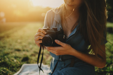 Closeup image of young hipster girl using vintage film camera while enjoying holidays in city park, beautiful woman photographer holding retro camera while walking in park, flare light effect