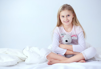 Little girl in the bedroom is sitting on the bed. Little girl is wearing a pajamas and sitting in bed