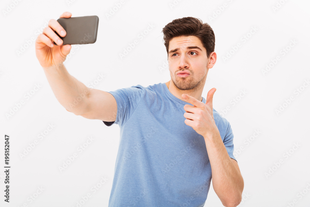 Sticker Image of attractive man with brown hair and bristle pointing index finger on camera while taking selfie on black mobile phone, isolated over white background